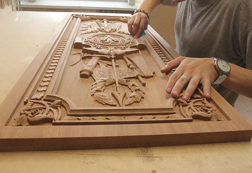 The craftsman polishes a carved relief with heraldic and weapon ornamentation, intended for wall interior decoration.