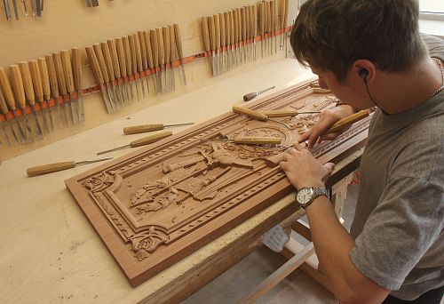 A master hand-carves a decorative relief panel with heraldic ornament on a wooden panel