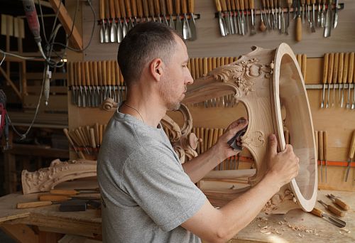 A craftsman in a gray t-shirt manually sands carved elements of an oak table in the workshop