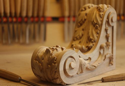 Hand-carved decorative oak element with elegant botanical ornament, set against a workshop backdrop with tools.