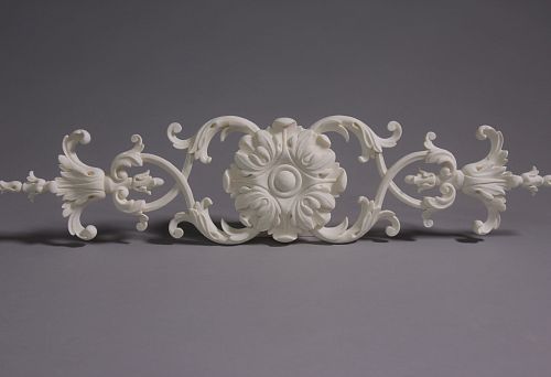 Classic molding decoration element with central rosette and symmetrical scrolls, executed in light tone on gray background.