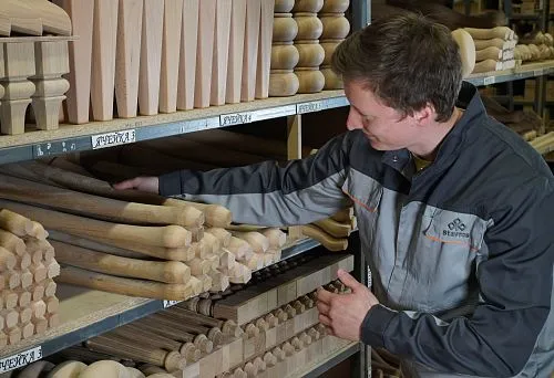 The craftsman selects wooden blanks on a shelf, demonstrating the variety of shapes and wood species for making chairs.