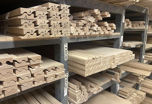 Long wooden sticks and carved blanks with decorative profiles, neatly arranged on metal shelves in a production workshop