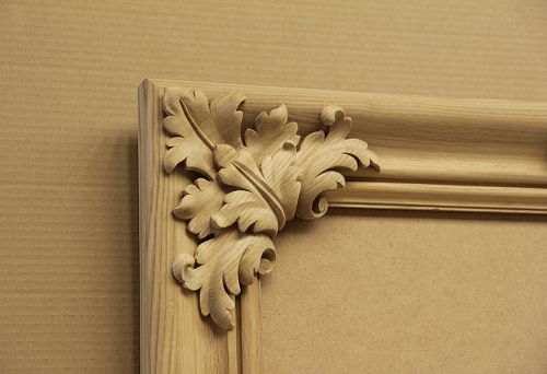 Corner piece of an unfinished carved wooden frame with a decorative oak leaf pattern on a kraft paper background.