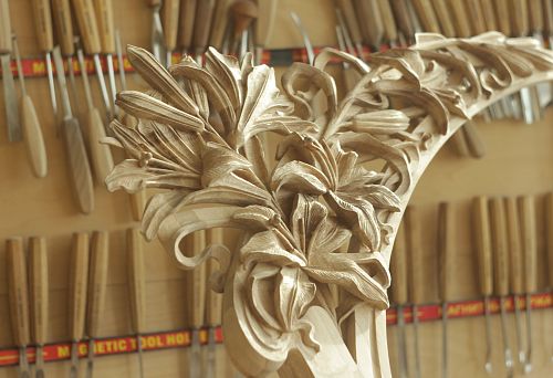 Hand-carved oak decorative element with large floral motifs, set against the backdrop of tools in the workshop.